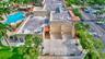 Stucco exterior facility featuring a concrete-surfaced outdoor racquetball court, adjacent swimming pool, palm-lined landscaping, and paved parking ar