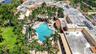Aerial view of a sprawling property featuring a custom-shaped swimming pool, extensive manicured lawn, abundant mature palm trees, and terracotta tile