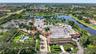 Aerial perspective of a multi-structure property featuring a swimming pool, expansive paved driveway, manicured landscaping with mature palm trees, an
