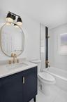Dark navy vanity with a stone countertop, brushed gold widespread faucet, oval brass-framed mirror, dual-shade wall lighting, and a bathtub with a ver