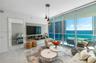 Floor-to-ceiling glass wall with ocean backdrop, private balcony, neutral large-format tile flooring, built-in wall-mounted media console, and triple-