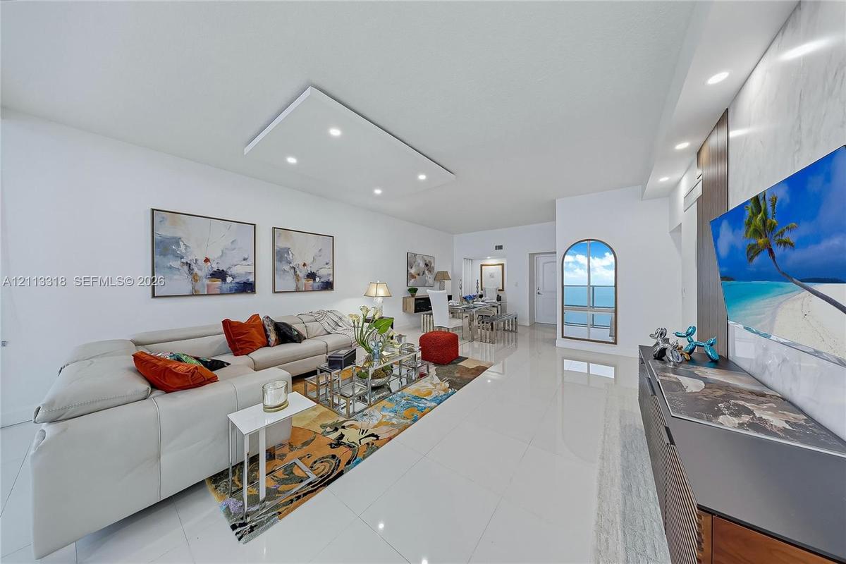 Open-concept living space featuring high-gloss tile flooring, recessed ceiling lighting, a custom stone-clad media wall, and an arched glass door