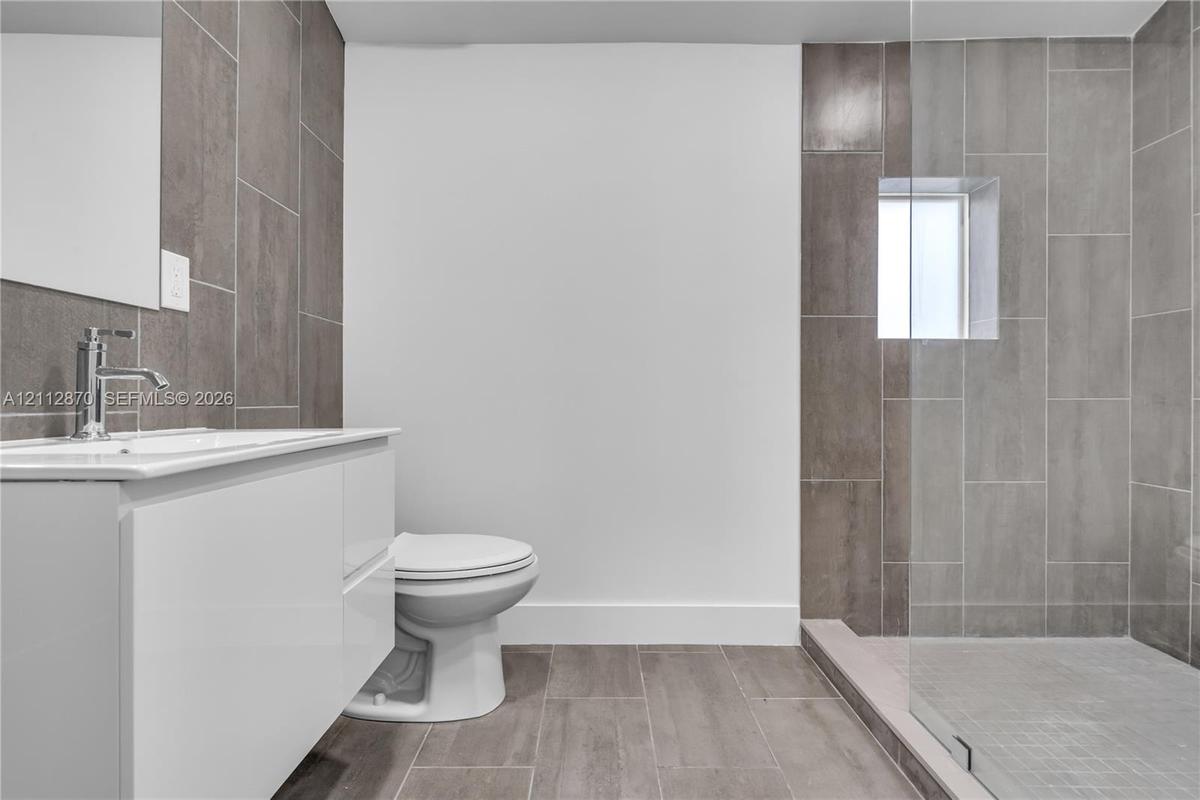 Bathroom featuring a white vanity cabinet, rectangular white countertop basin, chrome faucet, glass-enclosed shower, and large-format grey tile floori
