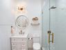 White vanity cabinet with a stone countertop, wall-mounted dual-bulb light fixture, circular bead-detail mirror, and a glass-enclosed shower with a ra