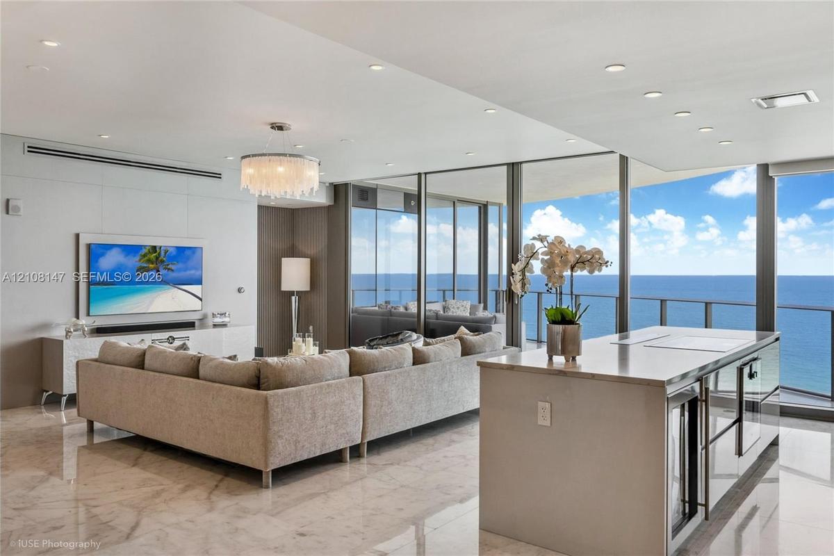 Open-concept interior featuring polished stone flooring, recessed lighting, a modern crystal chandelier, floor-to-ceiling glass panel walls, and a kit
