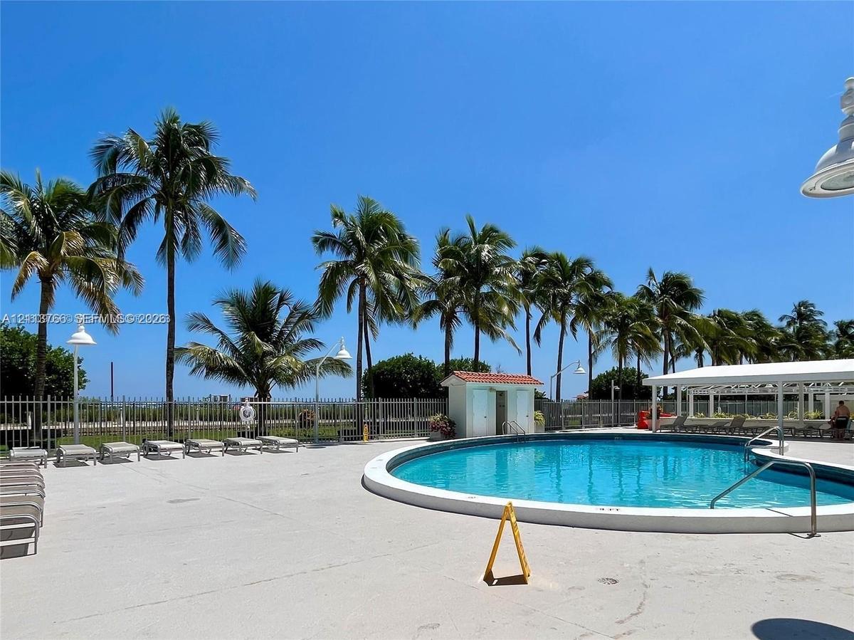 Circular swimming pool featuring a concrete deck, perimeter metal fencing, palm trees, and a covered pavilion