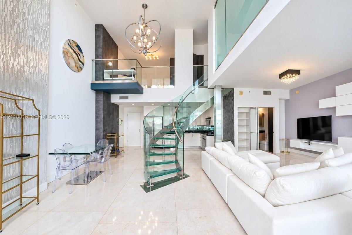 Double-height ceiling, glass-tread spiral staircase with metal railing, floor-to-ceiling textured accent wall, polished tile flooring, and glass mezza