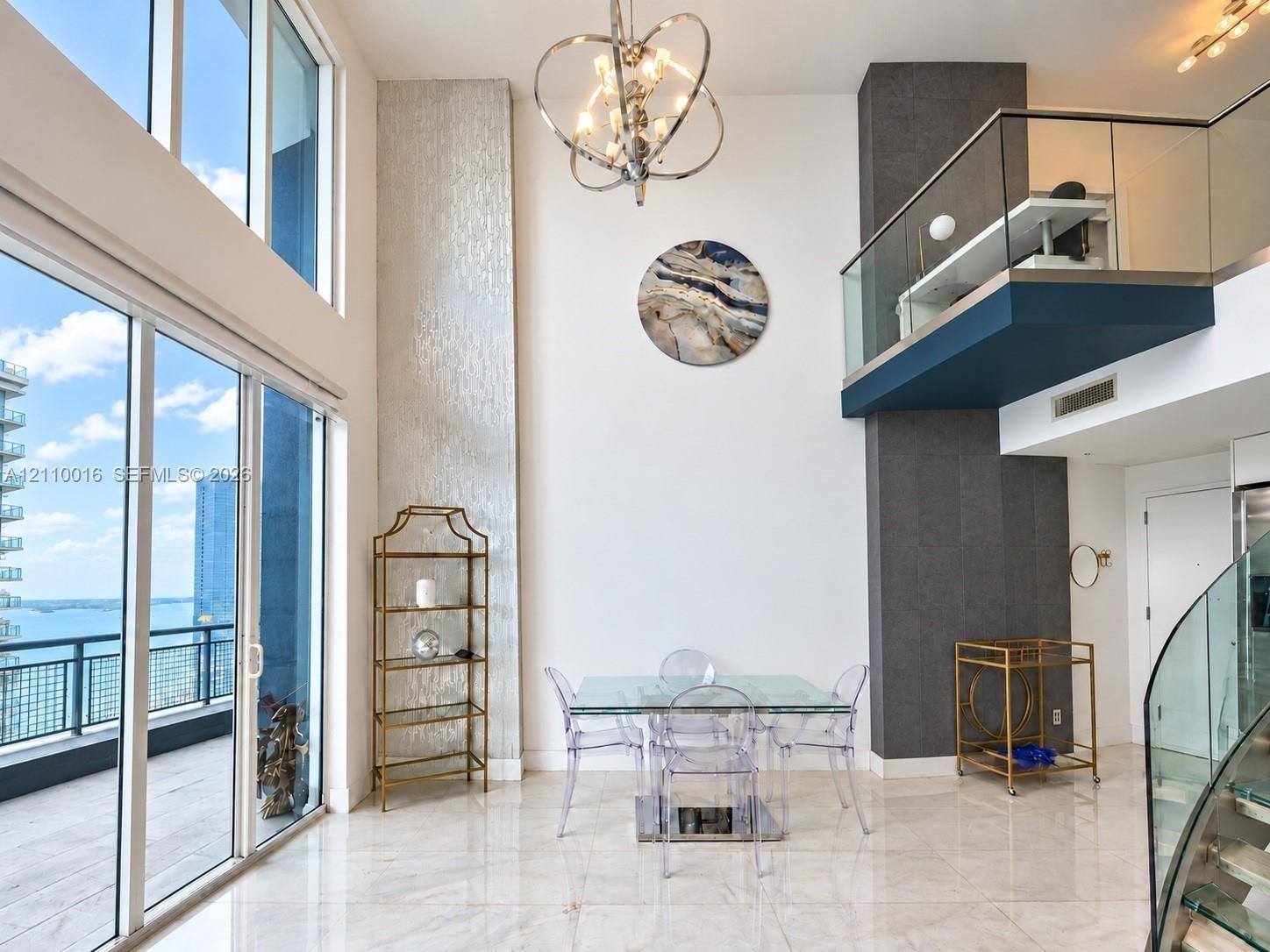 Double-height windows, balcony with glass railing, polished stone flooring, textured wall accent, and metal-finish chandelier