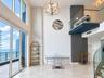 Double-height windows, balcony with glass railing, polished stone flooring, textured wall accent, and metal-finish chandelier