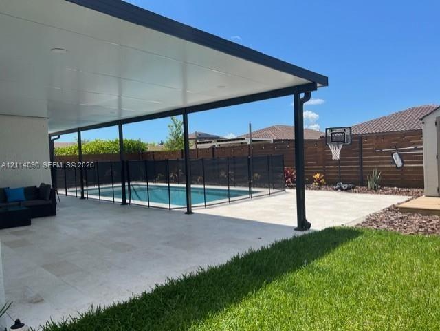 Covered patio featuring stone-finish pavers, recessed lighting, and a swimming pool with a black perimeter safety fence, set against horizontal slat p