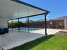 Covered patio featuring stone-finish pavers, recessed lighting, and a swimming pool with a black perimeter safety fence, set against horizontal slat p