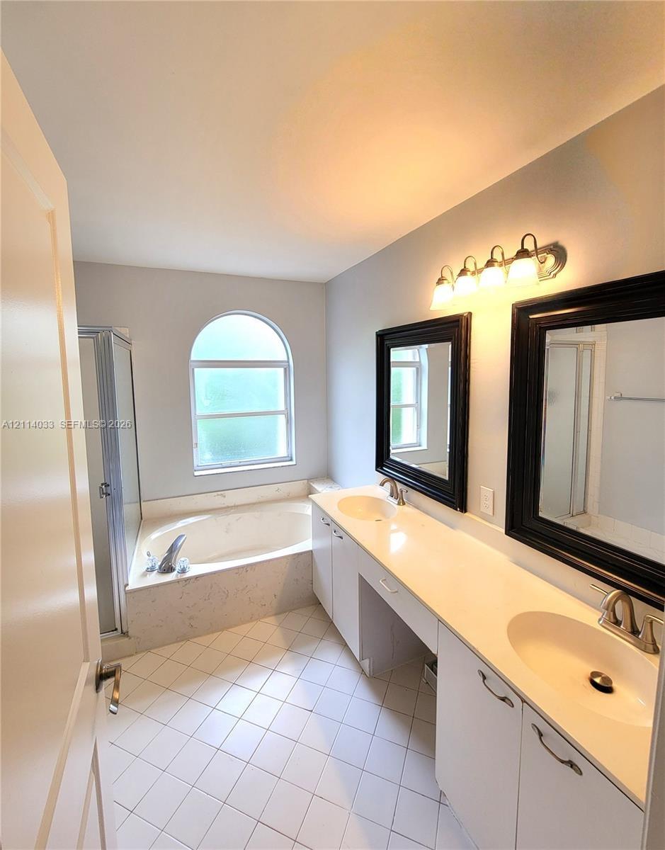 Bathroom featuring a dual-sink vanity with a solid-surface countertop, an arched window above a soaking tub with tile surround, a separate glass-enclo