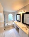 Bathroom featuring a dual-sink vanity with a solid-surface countertop, an arched window above a soaking tub with tile surround, a separate glass-enclo
