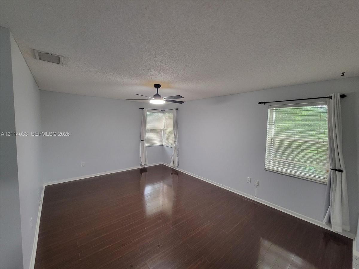 Spacious room featuring dark wood-finish flooring, a central ceiling fan with an integrated light fixture, dual-window corner configuration, and horiz