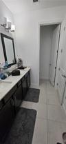 Double-sink vanity featuring dark cabinetry, light stone-finish countertops, three-light wall-mounted fixture, and oversized ceramic tile flooring