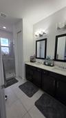 Dual vanity with dark cabinetry, light-toned countertop, framed mirrors, and glass-enclosed shower with tile flooring