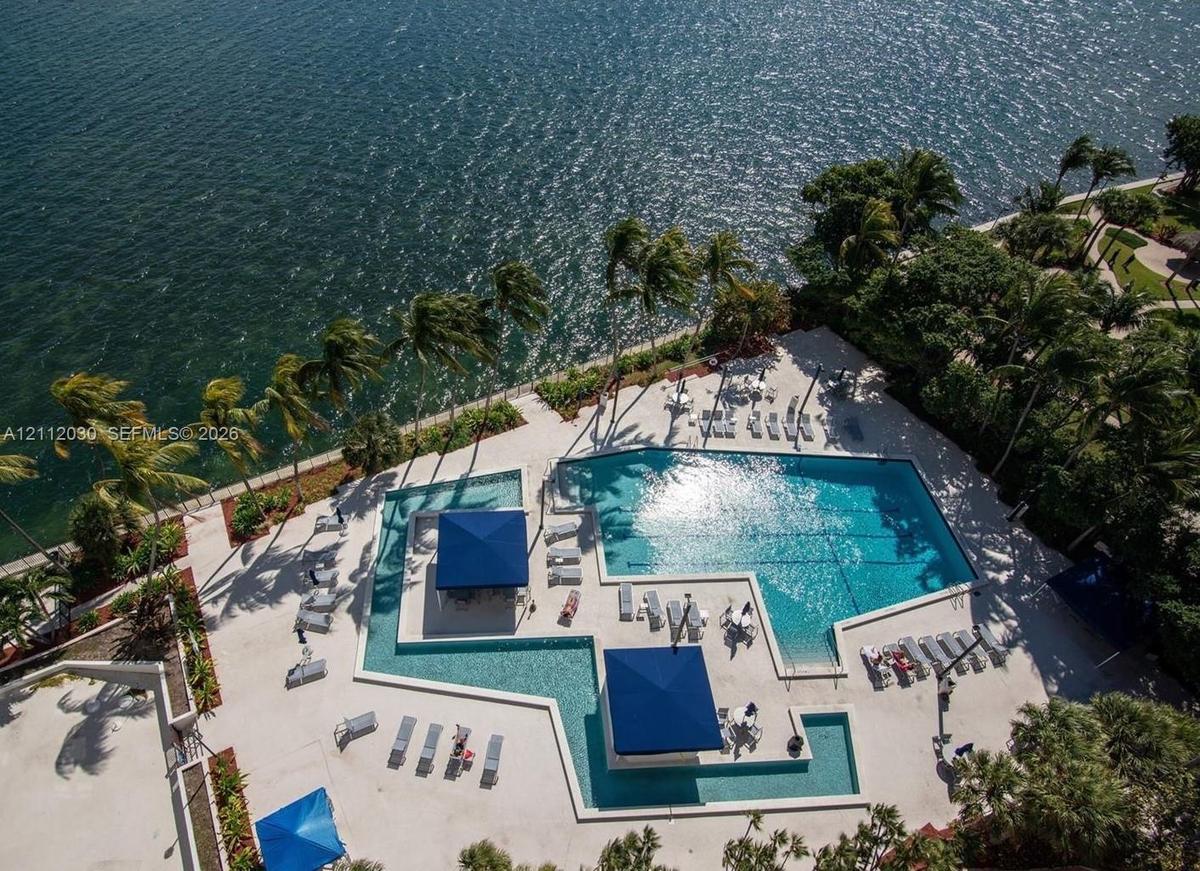 Waterfront pool complex featuring an expansive swimming area, integrated sun shelf, dual built-in shade structures, and surrounding paved decking