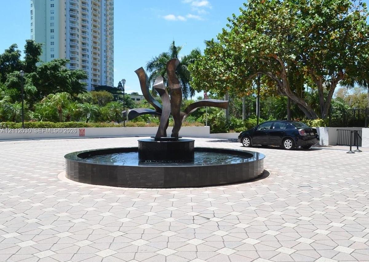 Circular stone-tiled motor court featuring a central dark stone fountain and abstract metal sculpture, framed by mature trees and landscaped stone per