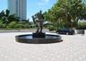 Circular stone-tiled motor court featuring a central dark stone fountain and abstract metal sculpture, framed by mature trees and landscaped stone per