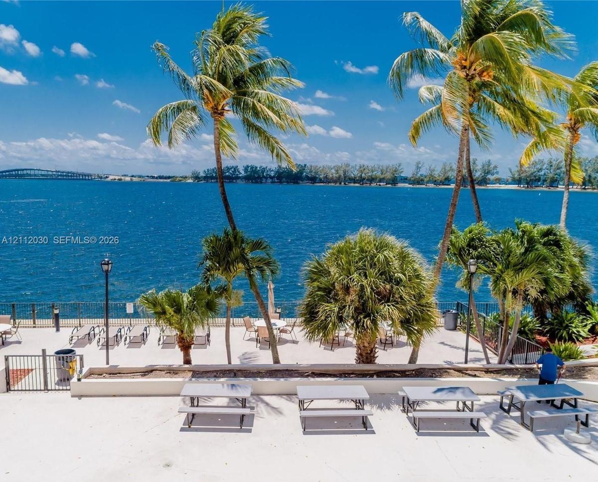 Waterfront patio featuring a concrete deck, metal perimeter fencing, integrated lighting, and mature palm trees