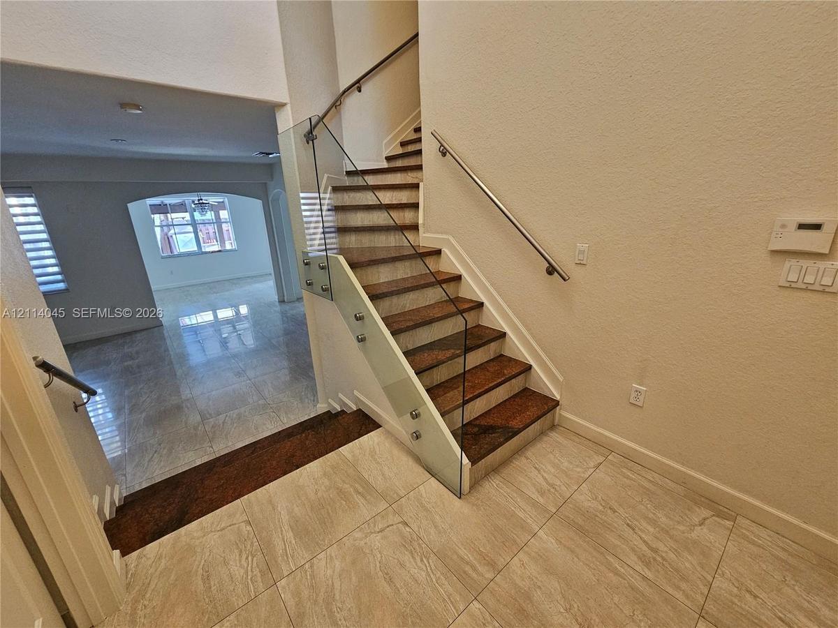 Staircase featuring wood-finish treads, glass panel railing with metallic standoffs, and a wall-mounted metal handrail, complemented by neutral tile f
