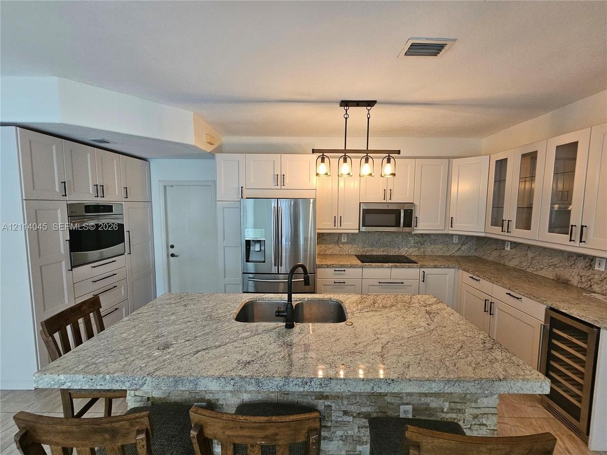 Kitchen featuring white cabinetry, stone countertops, a central island with an integrated sink, stainless steel appliances, and a built-in wine cooler