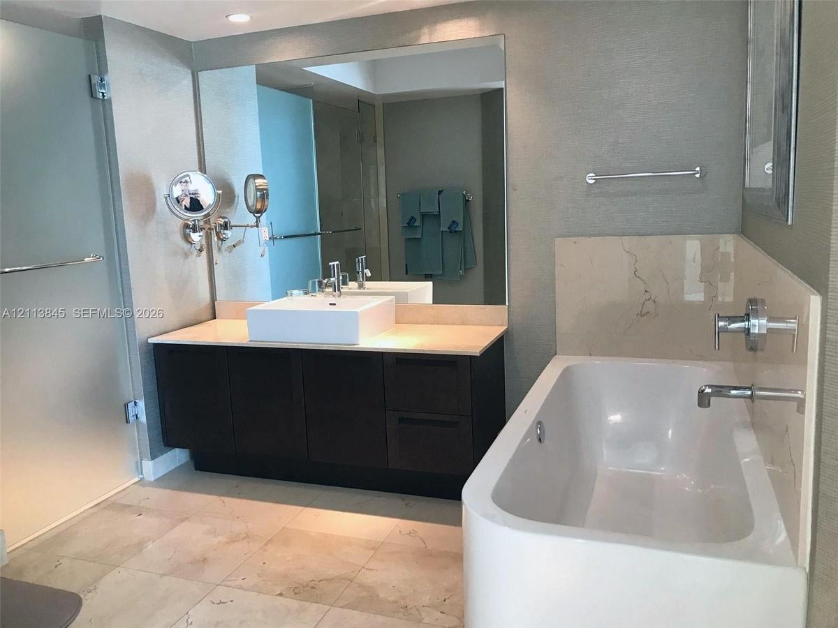 Dark wood-finish vanity with a rectangular vessel sink and countertop, large wall-mounted mirror, soaking tub with polished chrome fixtures, and large