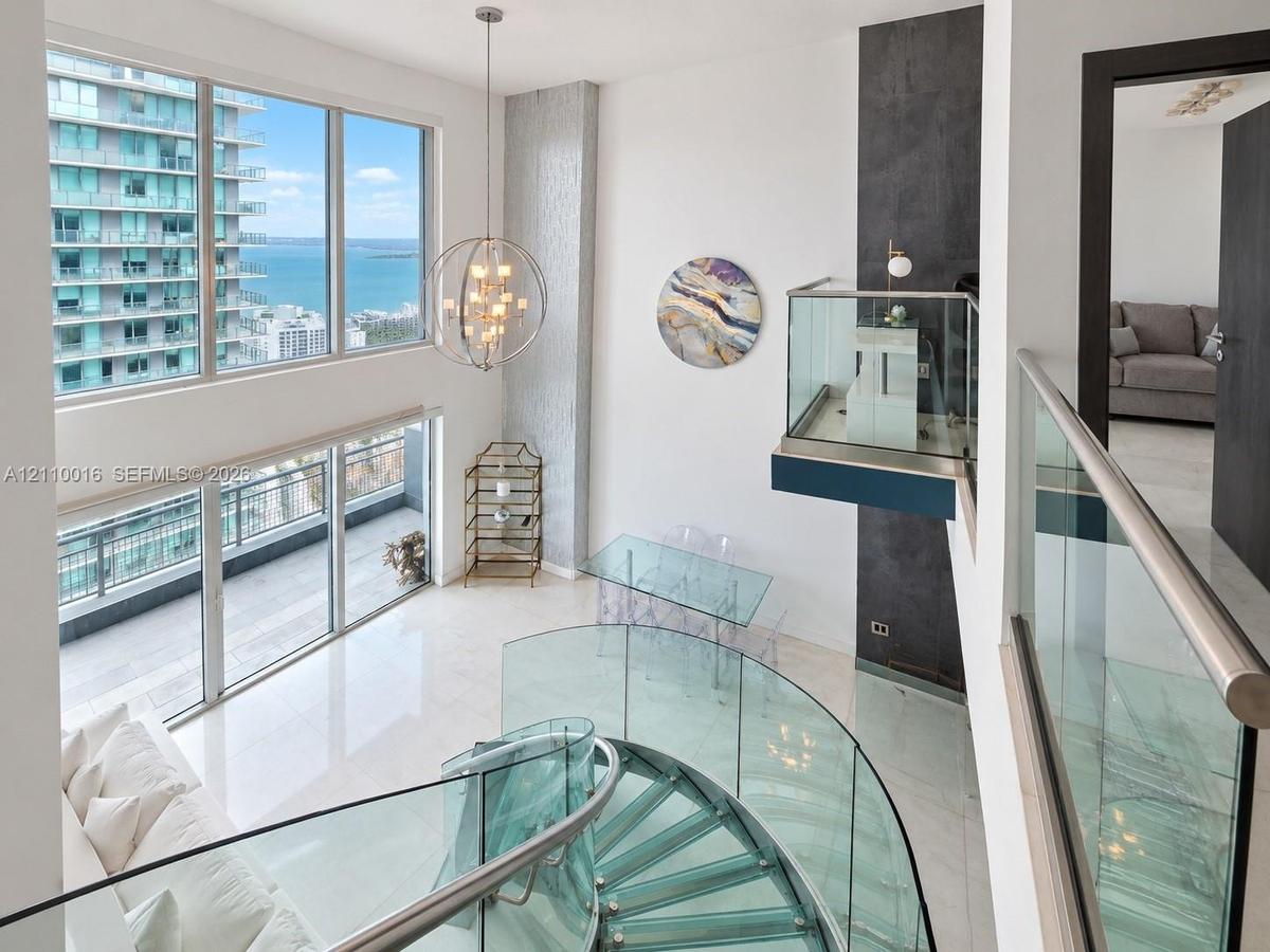 Double-height living area featuring floor-to-ceiling windows, polished stone flooring, custom glass-panel staircase with metal railing, and contempora