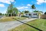 Single-story exterior featuring a light-toned stucco facade, concrete driveway, expansive grass lawn, mature palm trees, and a wooden privacy fence