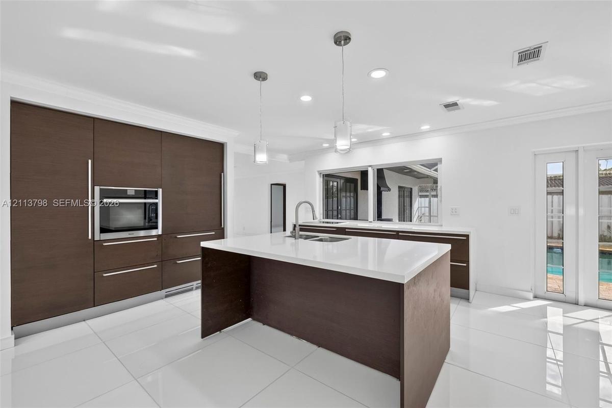 Kitchen featuring dark wood-finish cabinetry, solid-surface countertops, a center island with a built-in sink, stainless steel wall oven, and polished