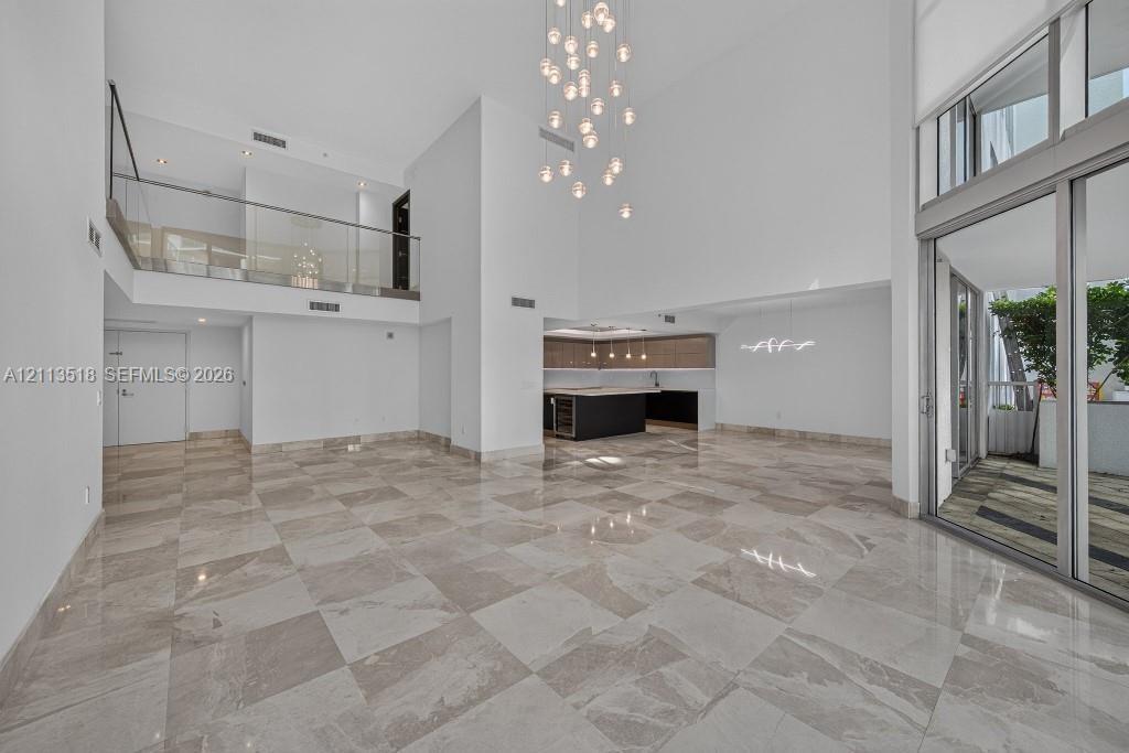Open-concept interior featuring double-height ceilings, stone-finish tile flooring, a glass-panel mezzanine railing, multi-light pendant chandelier, a