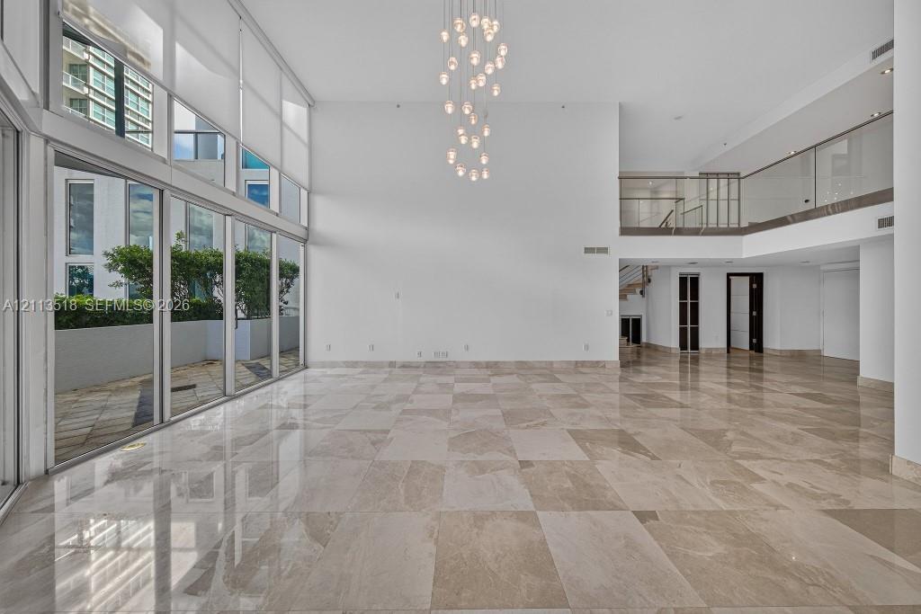 Double-height ceilings, floor-to-ceiling glass wall, polished stone flooring, multi-tiered pendant light fixture, and second-level mezzanine with glas