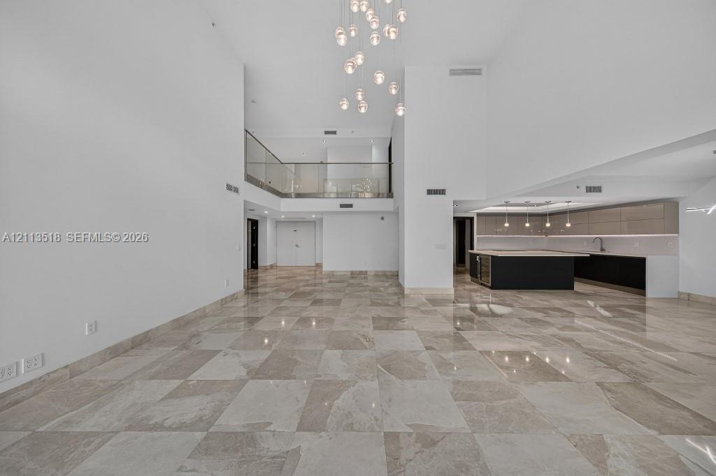 Expansive interior featuring high ceilings, stone flooring, a multi-tiered glass pendant chandelier, a glass-railed mezzanine, and kitchen cabinetry w