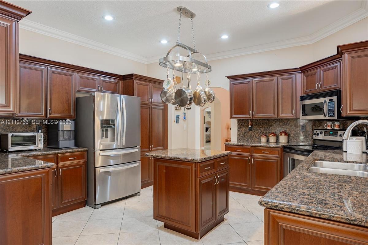 Kitchen featuring stained wood cabinetry, stone-surface countertops, tile flooring, stainless steel appliances, and a suspended metal pot rack
