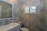 Bathroom featuring patterned wall covering, white vanity with rectangular sink, polished chrome faucet, framed mirror, and white plantation shutters