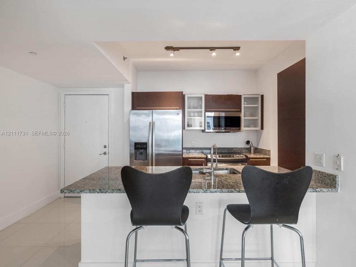 Kitchen featuring a stone countertop peninsula, stainless steel refrigerator and built-in microwave, flat-panel wood-finish cabinetry with glass-front