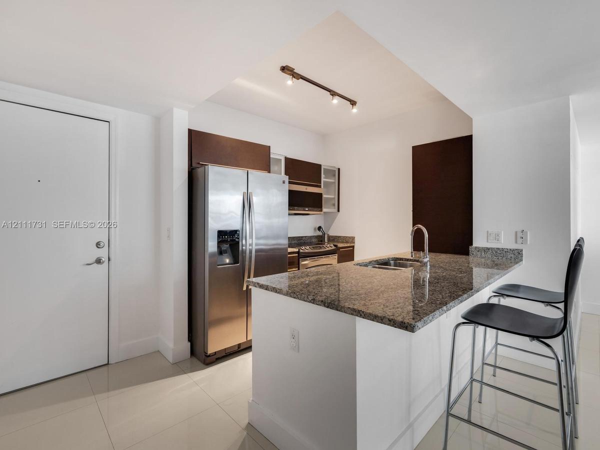 Kitchen featuring stone-surfaced countertops, a center island with an integrated sink, dark wood-finish cabinetry, stainless steel appliances, and tra