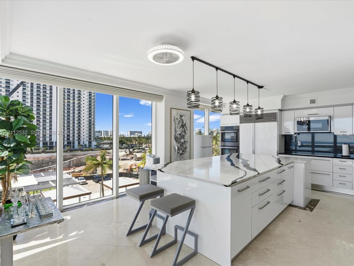 Kitchen featuring high-gloss cabinetry, stone-topped center island with waterfall edge, built-in double wall ovens, five-light linear pendant fixture,