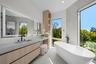 Floating wood-finish vanity with integrated sink, expansive LED-backlit mirror, freestanding soaking tub with floor-mounted faucet, and floor-to-ceili