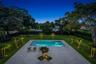 In-ground swimming pool featuring a tanning ledge, light-toned stone patio, manicured lawn, perimeter fencing, and established palms