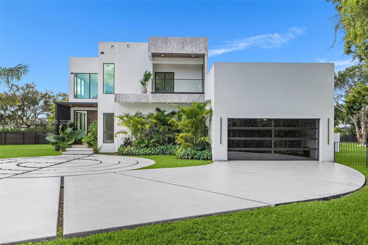 Modern two-story exterior featuring smooth stucco siding, a second-level balcony with glass railing, tiered concrete driveway with decorative inlay, a