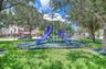 Community park featuring a central playground structure with dual slides, a canopy cover, manicured lawn, and mature canopy trees