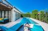 Rear exterior featuring a covered patio, rectangular in-ground pool with integrated spa, light-toned stone decking, and mature privacy hedges