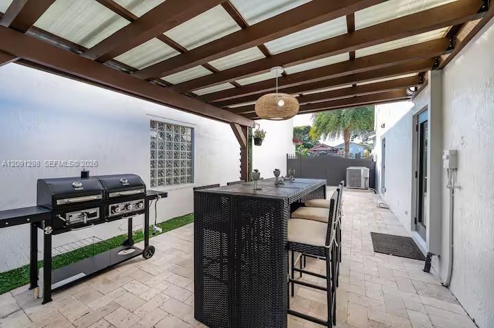 Covered patio featuring a wood-beam pergola, paver flooring, a glass block window, and a hanging pendant light fixture