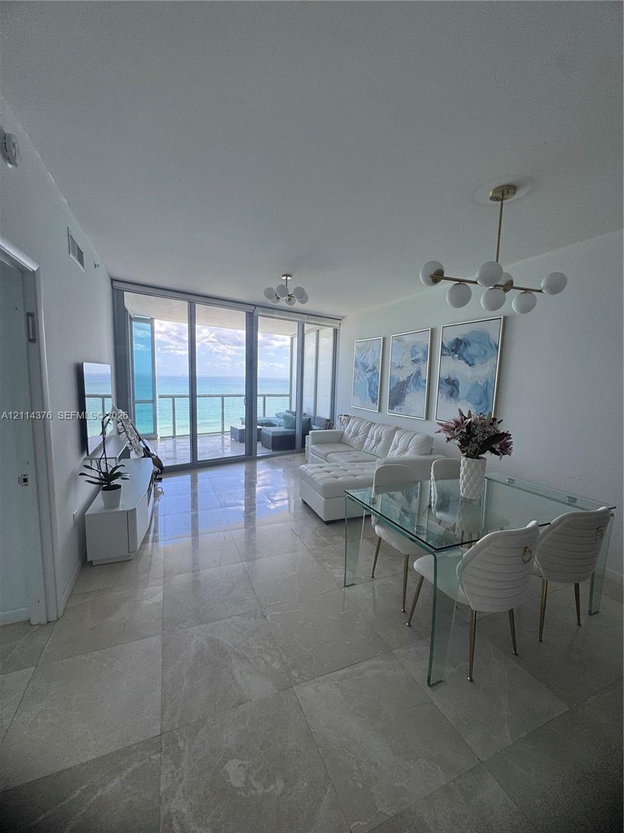 Open-concept living space featuring floor-to-ceiling glass panels, large-format tile flooring, multi-globe brass-finish chandeliers, and a private bal