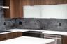 Kitchen featuring wood-finish cabinetry, matte black gooseneck faucet, white solid-surface countertops, textured dark stone backsplash, and frosted gl