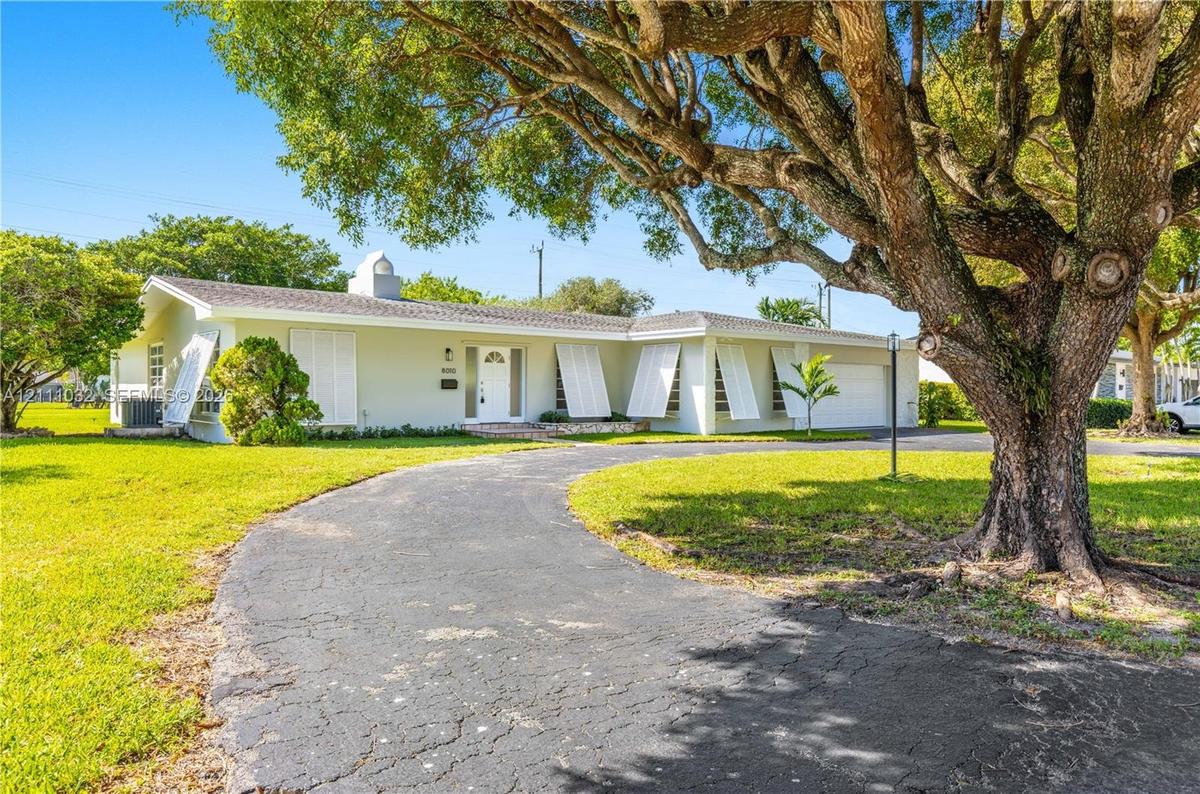 Single-story residence featuring a curved paved driveway, exterior shutters, attached garage, and mature landscaping on a spacious lot