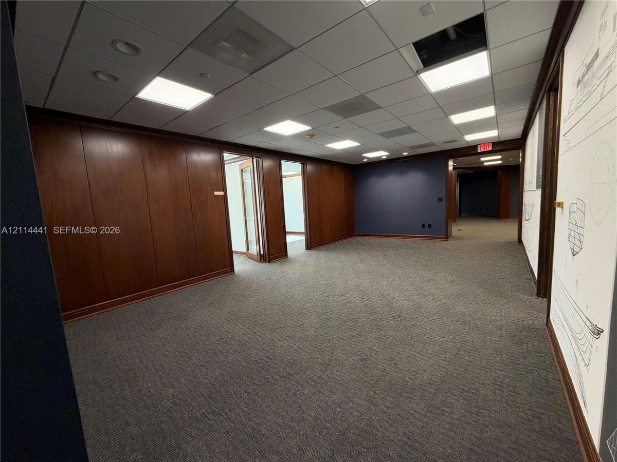 Spacious interior featuring full-height wood paneling, drop ceiling with recessed panel lighting, carpeted flooring, and dark-toned accent walls