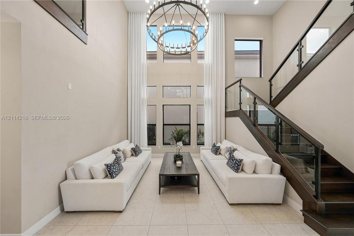 Two-story height ceiling with expansive windows, custom iron-frame chandelier, and staircase featuring dark wood finishes and glass railing panels