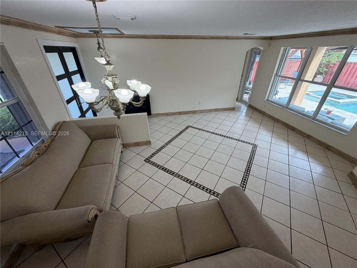 Living area featuring large-format ceramic tile flooring with decorative inlay border, ornate multi-arm chandelier, crown molding, and expansive windo